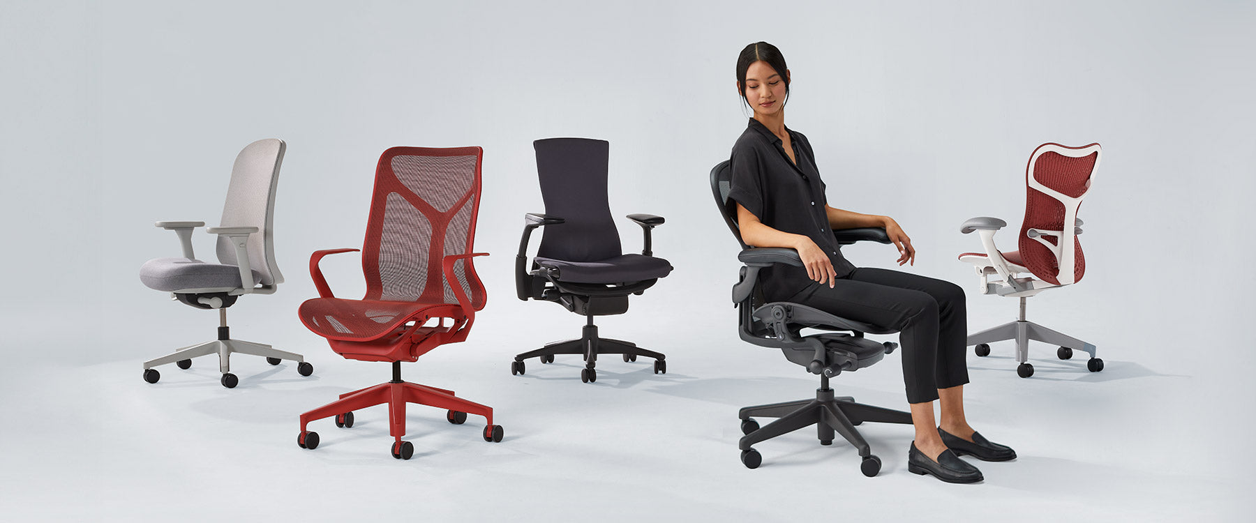 Photo of Herman Miller chairs lined up including Verus mineral white & grey, Cosm mid back in canyon red, Embody black, Aeron graphite with lady sat in and Mirra 2 in cordovan red