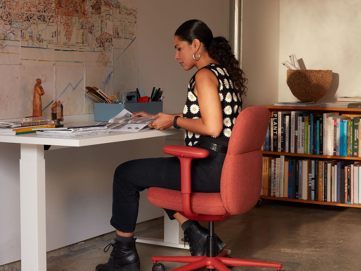 Anooshey Rahim sat working on a Asari mid back office chair in canyon red at a Nevi sit-stand desk