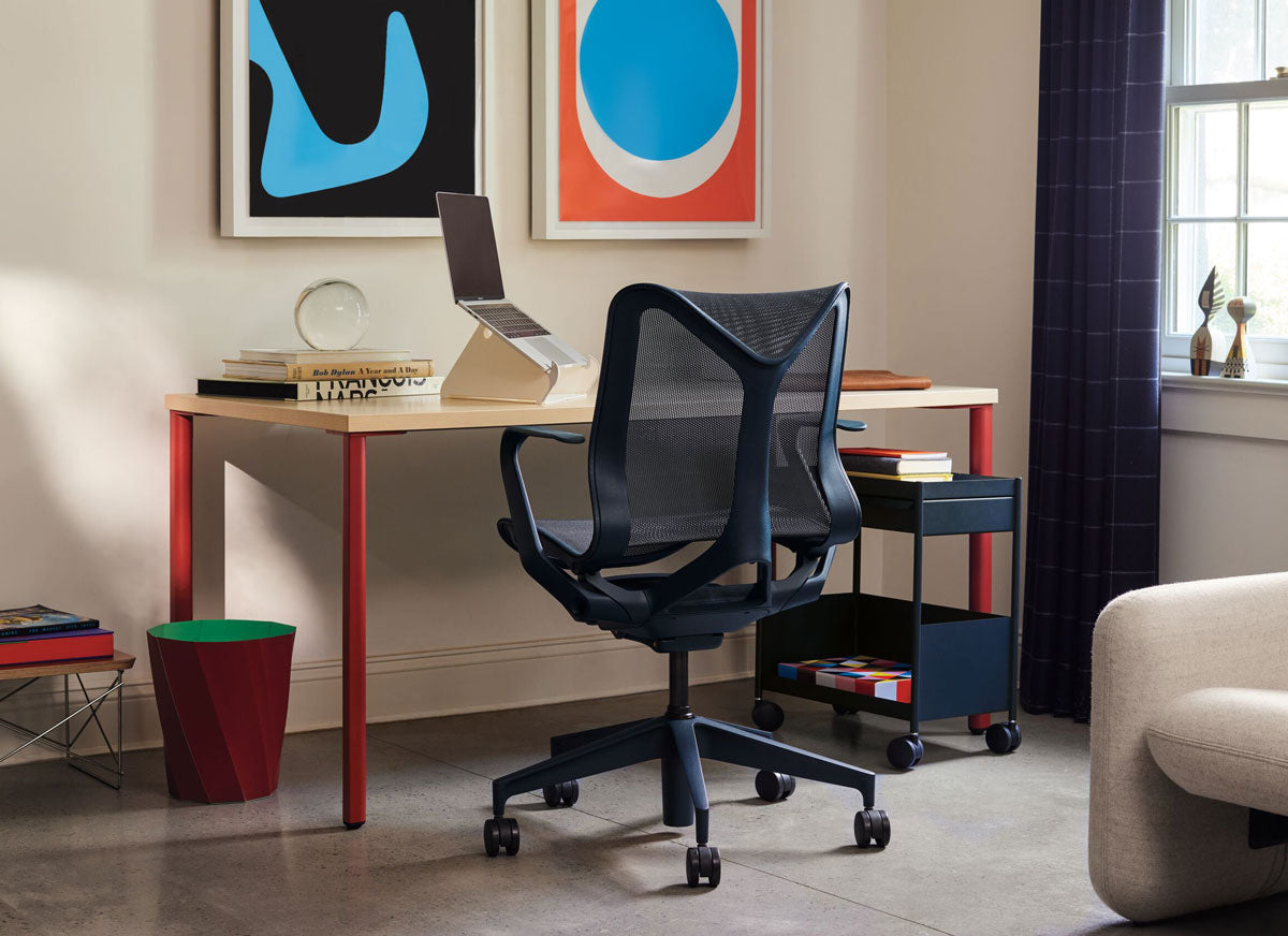 A home office setup featuring a wooden-topped OE1 desk with red legs and a nightfall blue Herman Miller Cosm ergonomic desk chair.