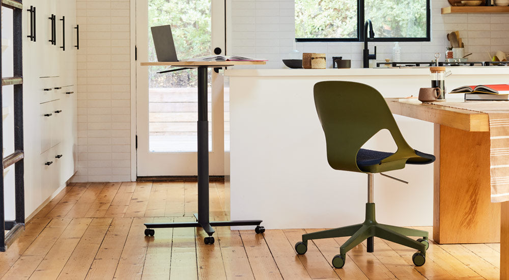 A black and oak Passport table by Herman Miller positioned near a white kitchen island, with a laptop and book. The image also features a green and black Zeph chair by Herman Miller positioned at a wooden dining table.