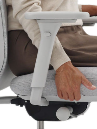 Close-up side view of a person sitting on a grey Lino office chair by Herman Miller. Dressed in a beige jumper and brown trousers, they are using the adjustable seat depth feature.