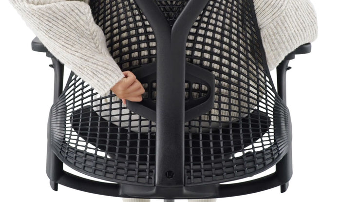 Close-up rear view of a person sitting in a black Sayl office chair by Herman Miller, reaching behind to move the height-adjustable lumbar support.