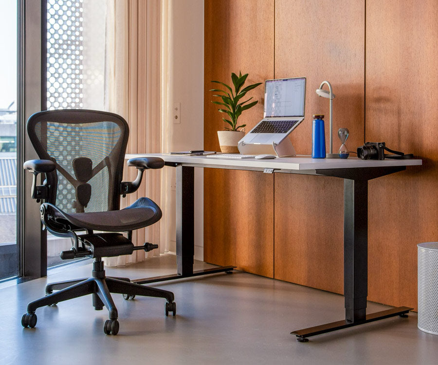 Home office setting showing Nevi sit-stand desk in a seated position with black legs and white top with Aeron office chair in graphite