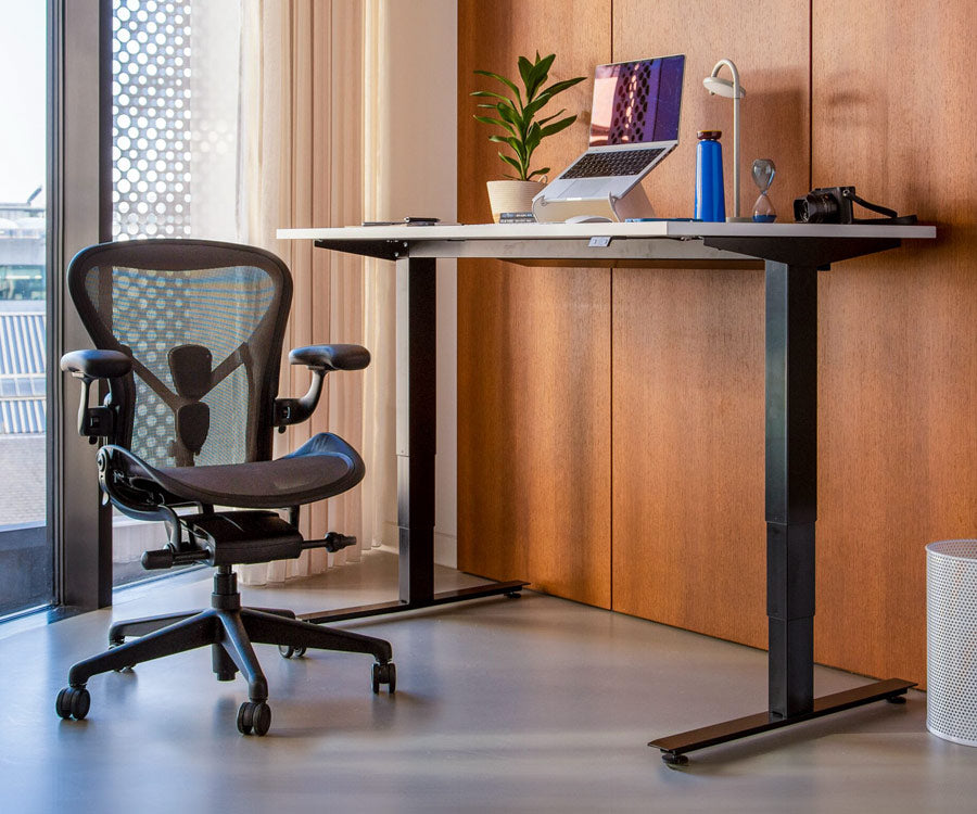 Home office setting showing Nevi sit-stand desk in a standing position with black legs and white top alongside an Aeron office chair in graphite in background
