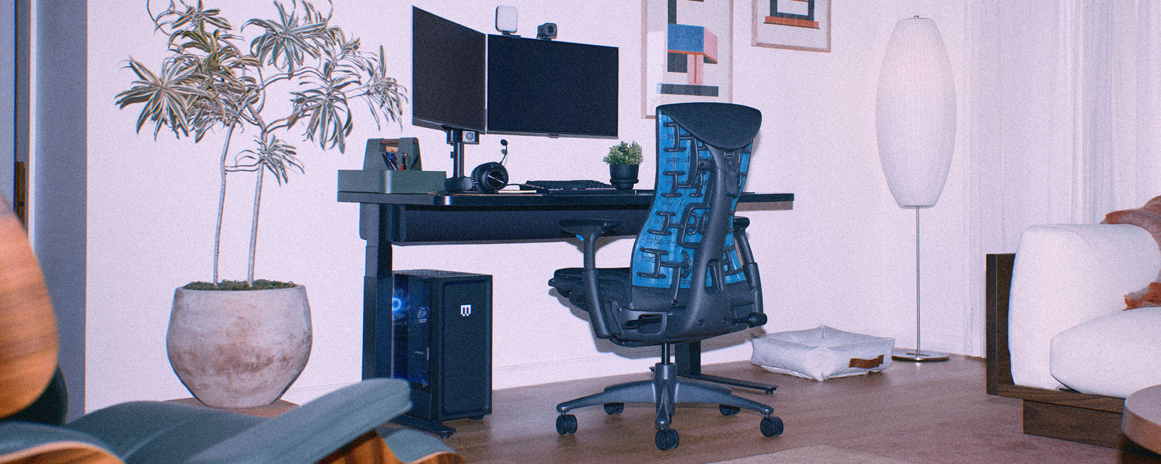 A grainy image of a black and cyan blue Herman Miller Embody gaming chair as part of a home PC gaming setup including a Nevi sit-stand gaming desk.