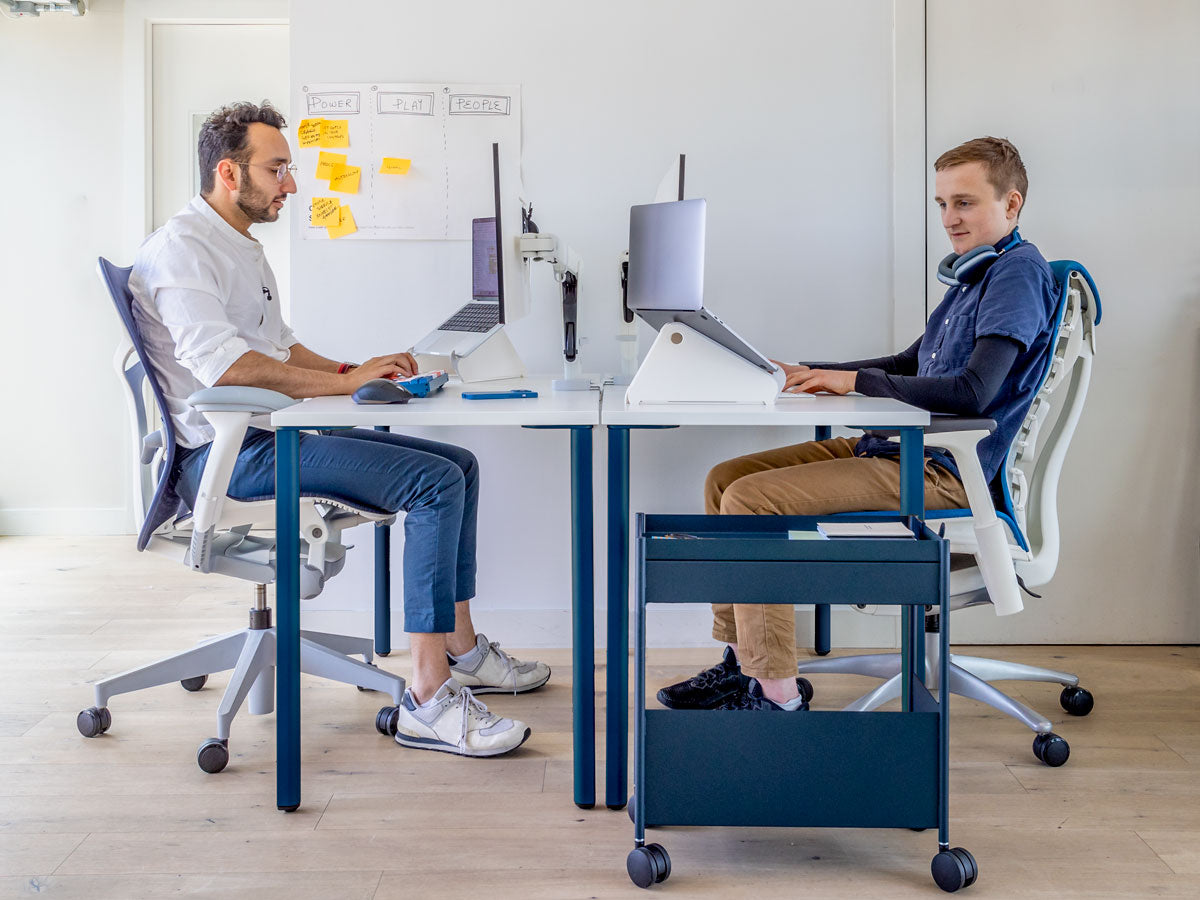 Photo of 2 men sat at desk opposite each showing that when you sit properly your elbows are level with top of desk. Features OE1 desk with blue legs and white top along with Mirra 2 in mineral white & blue and Embody in white