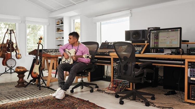 Photo of Rostam, muscian, producer and composer, in his music studio playing guitar sat in Aeron office chair in graphite