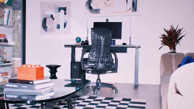 A black Herman Miller Embody ergonomic gaming chair as part of a PC gaming setup including a Nevi sit-stand desk in a bright living space.