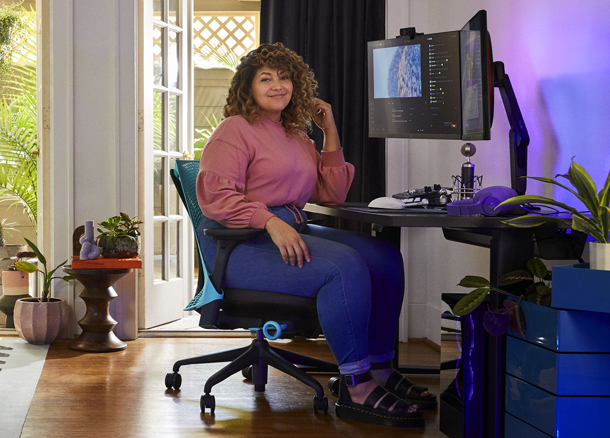 A woman sitting in a blue and black Sayl gaming chair by Herman Miller, positioned behind a black Nevi desk with a black Ollin monitor arm and screen. Set in an indoor space with wooden floors, white walls and indoor plants.