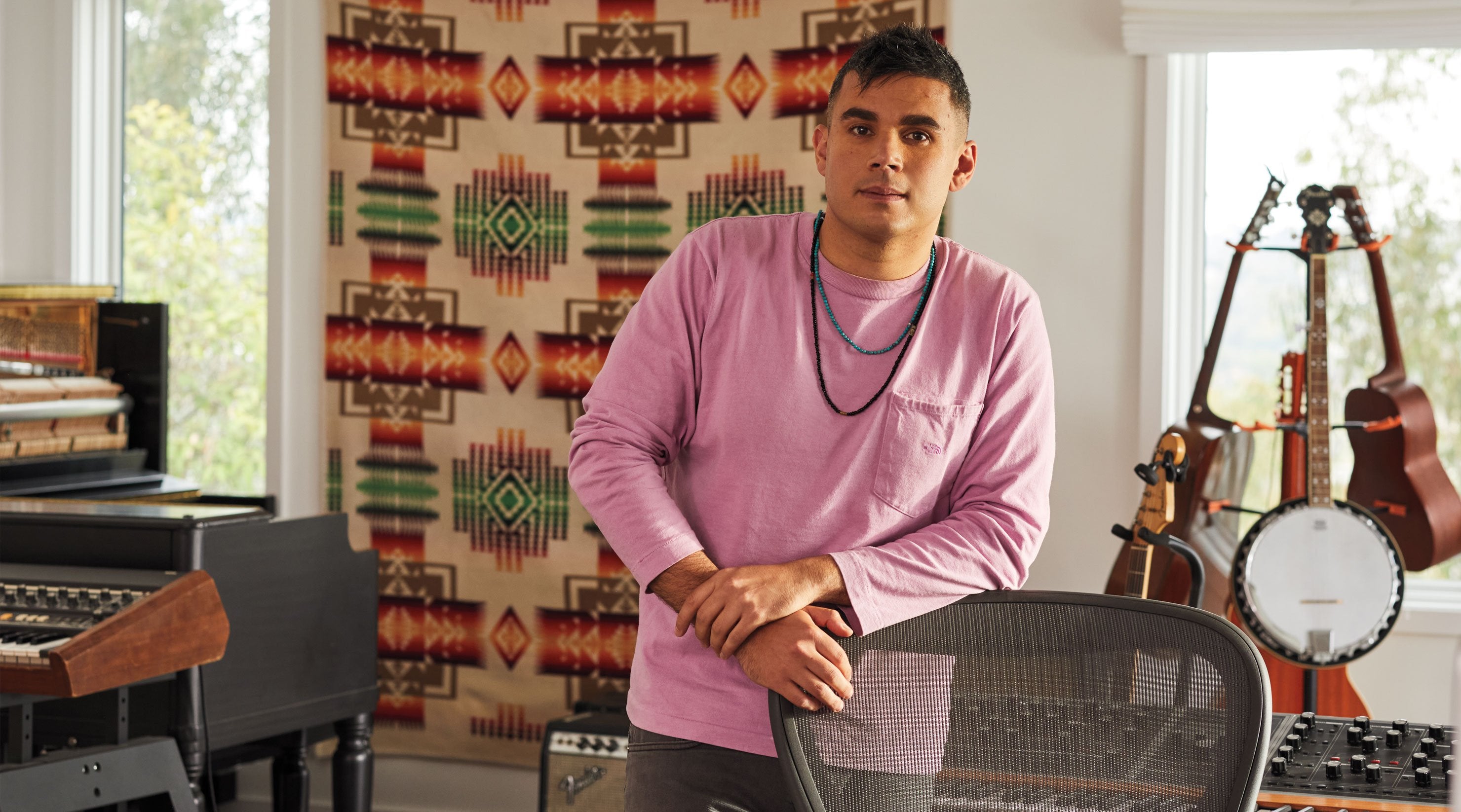 Photo of Rostam, muscian, producer and composer, stood leaning on Aeron office chair in graphite with musical instruments in the background