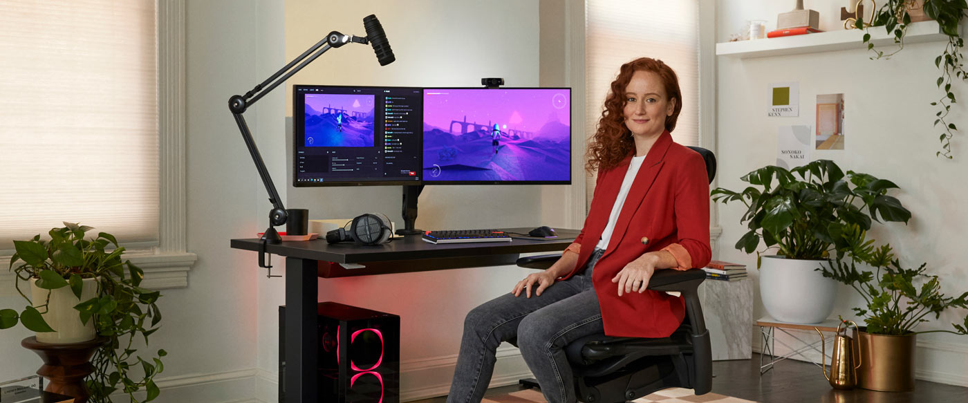 Photo of Dr Lindsey Migliore sat in a Embody gaming chair facing camera with Nevi gaming desk in black with ollin monitor arms