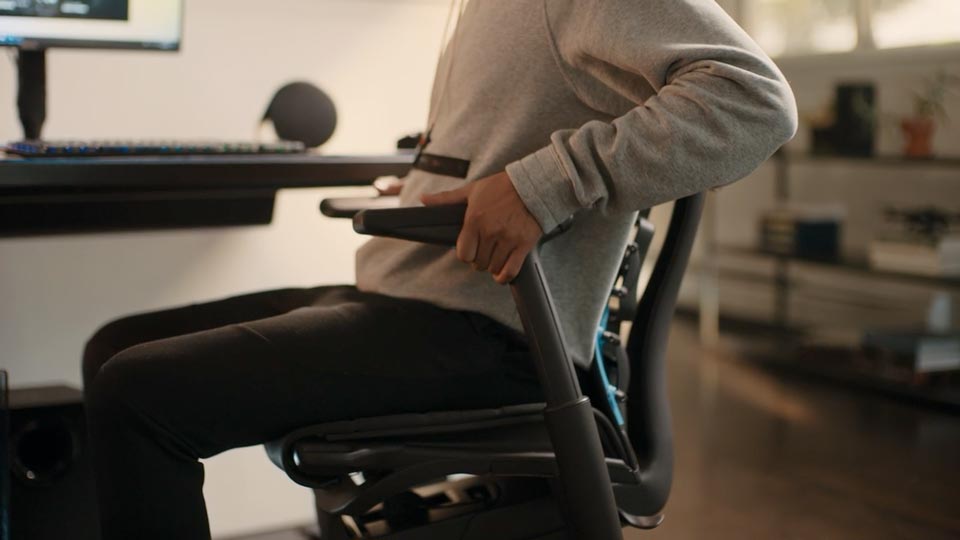 A video of a person sitting in a black and blue Embody gaming chair by Herman Miller, reaching beside the chair to adjust the arms.