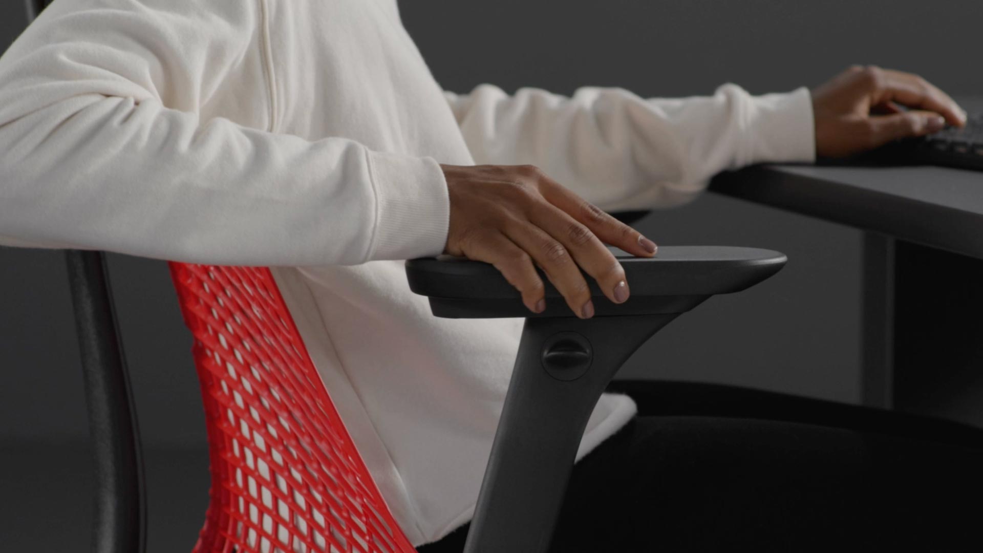 A video of a person sitting in a red and black Sayl gaming chair by Herman Miller, reaching beside the chair to adjust the arms.