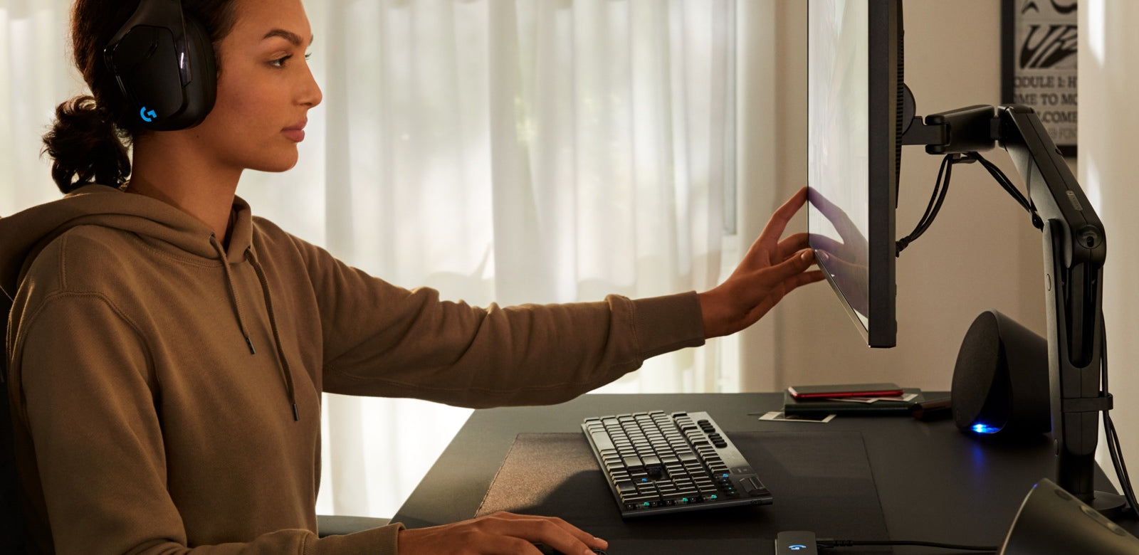 A woman sat at a black Nevi gaming desk with a black Ollin gaming monitor arm by Herman Miller. She is wearing Logitech headphones and moving the computer screen with one of her hands.