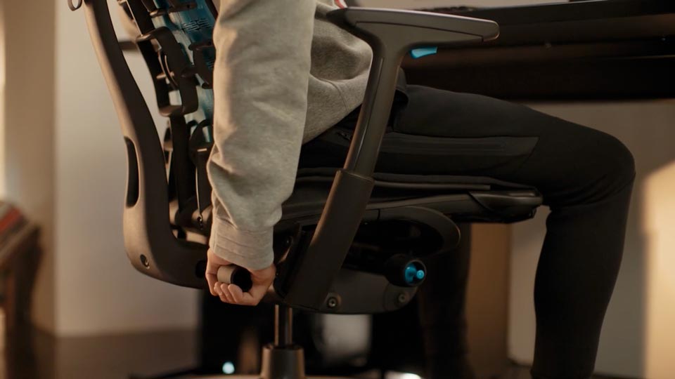 A video of a person sitting in a black and blue Embody gaming chair by Herman Miller, reaching behind the chair to adjust the back support.