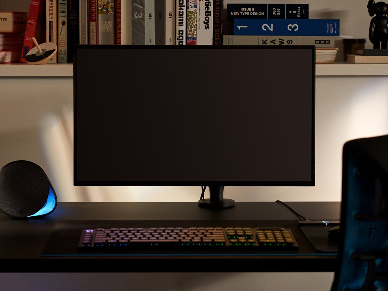 Front view of a black Ollin monitor arm holding a computer screen and attached to a black Nevi gaming desk by Herman Miller. Set in a games room or home office, with a bookshelf, keyboard and mouse.