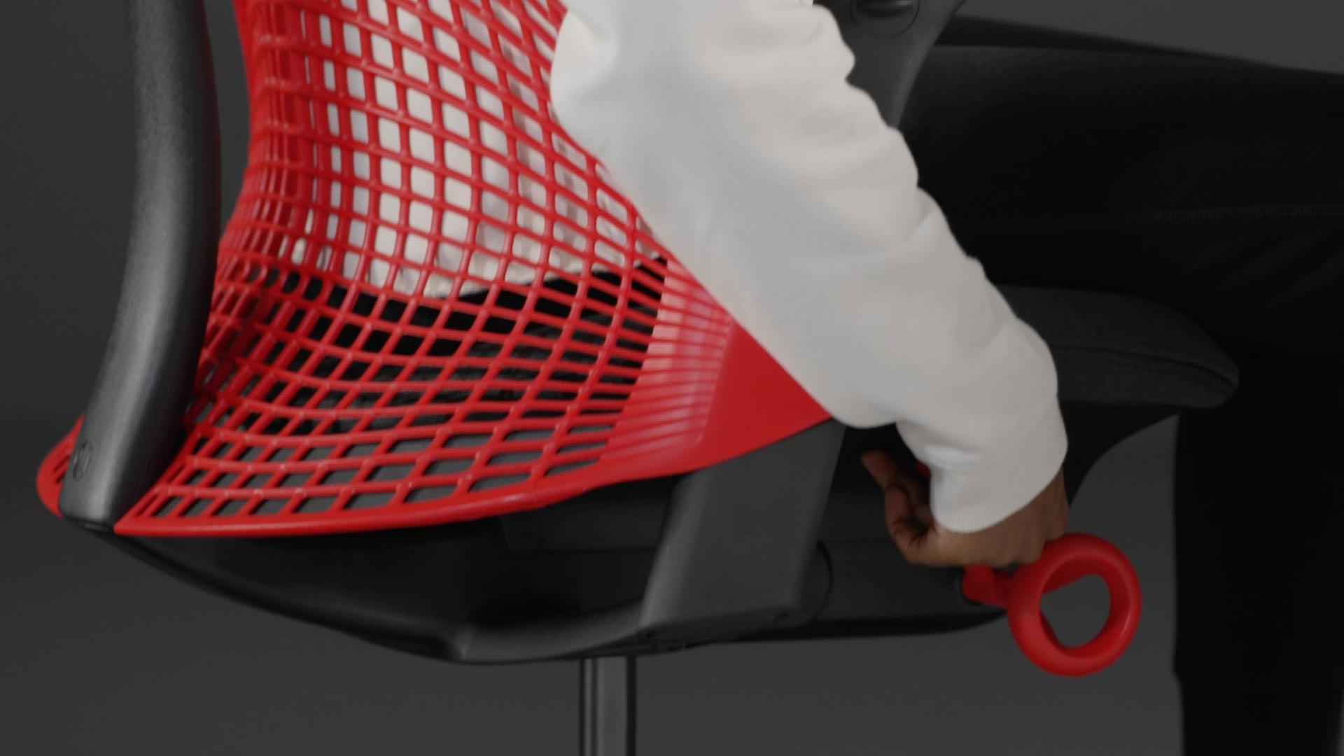 A video of a person sitting in a red and black Sayl gaming chair by Herman Miller, reaching beside the chair to adjust the seat height.