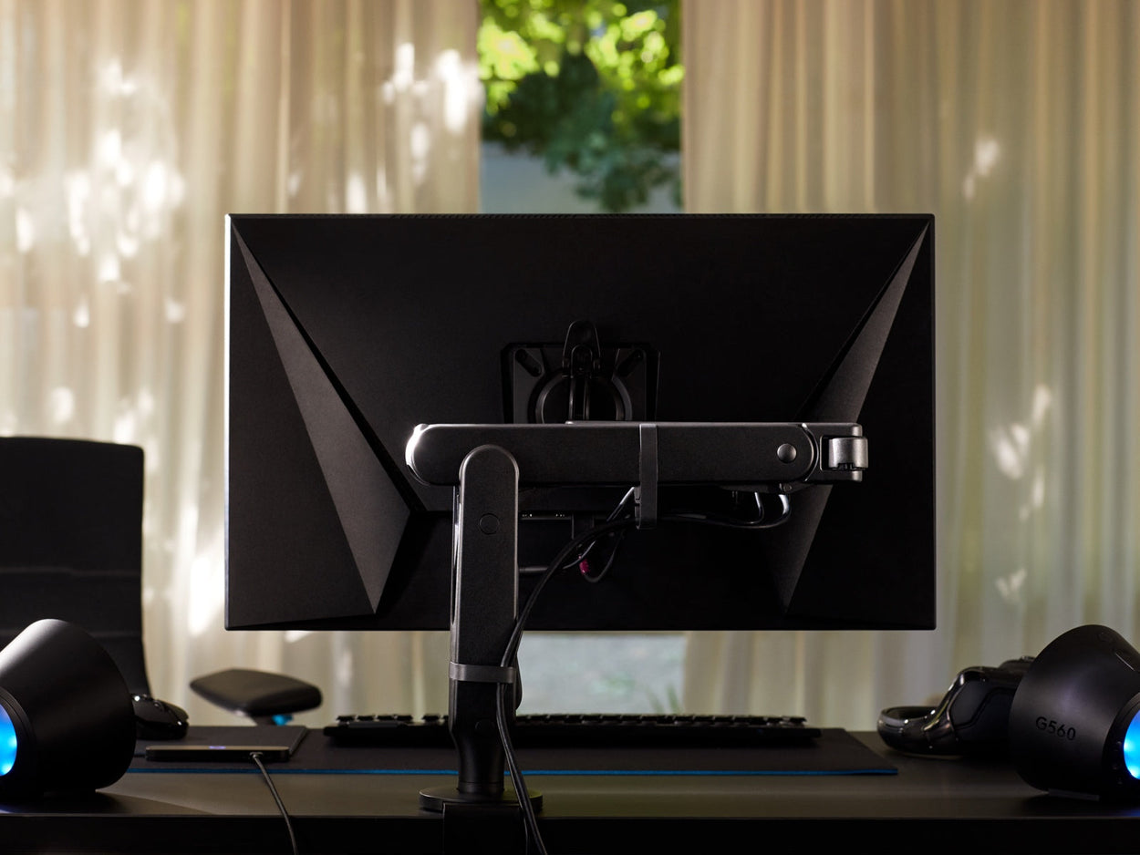 Rear view of a black Ollin monitor arm holding a computer screen and attached to a black Nevi gaming desk by Herman Miller. Set in a games room or home office, with lights, keyboard and mouse.