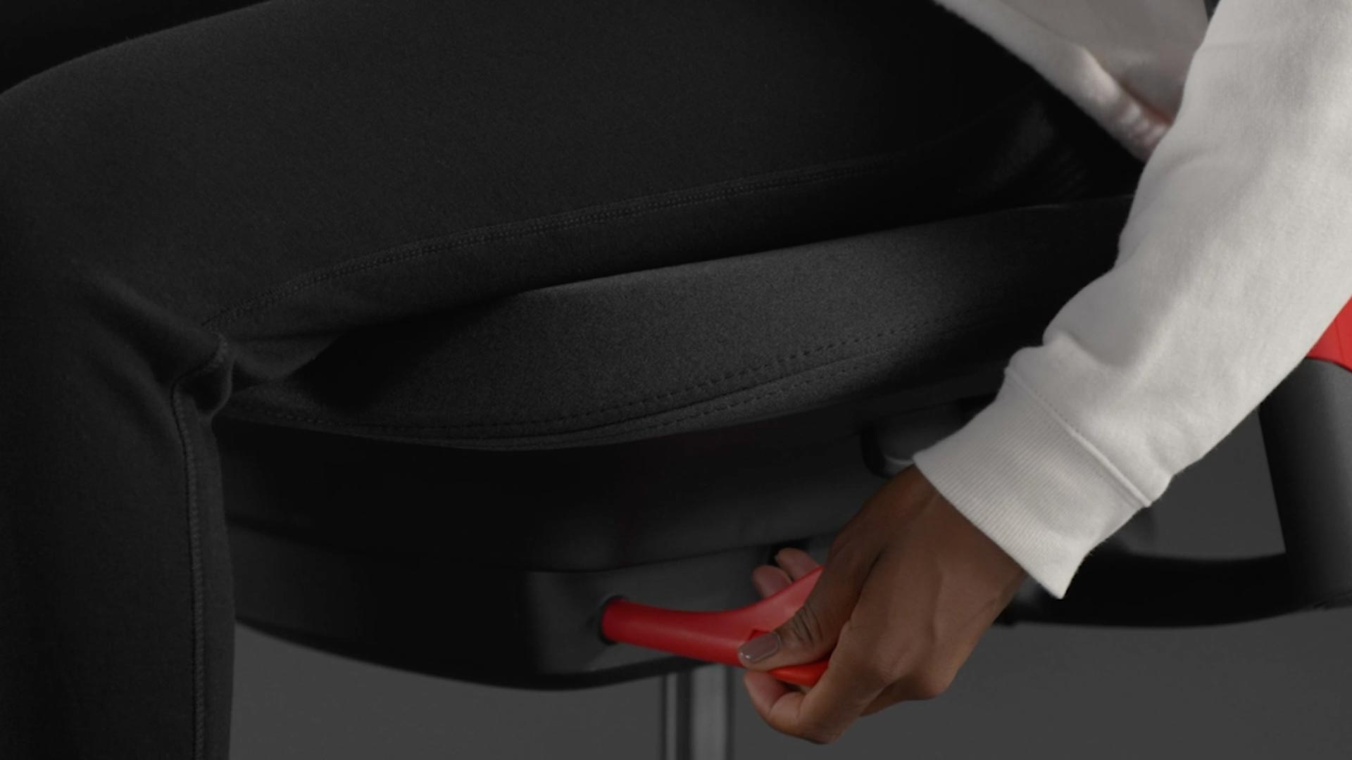 A video of a person sitting in a red and black Sayl gaming chair by Herman Miller, reaching beside the chair to adjust the forward tilt.