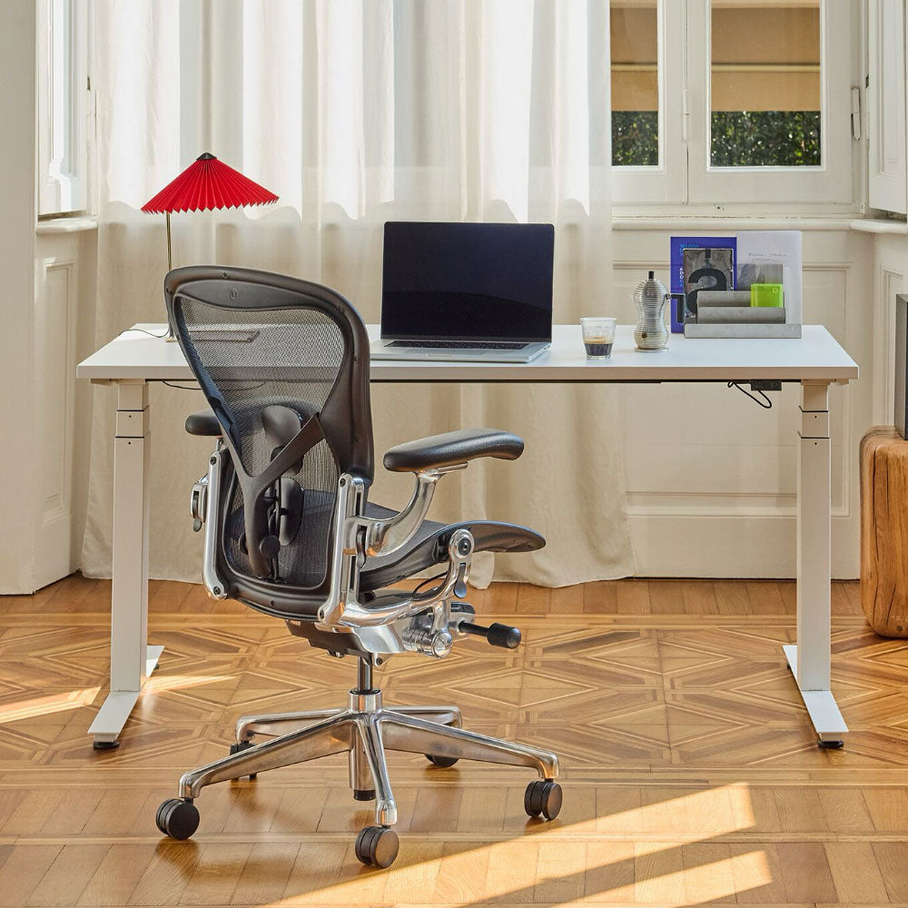 A warm home office with natural materials featuring a graphite and chrome Herman Miller Aeron ergonomic chair and a white Nevi sit-stand desk.