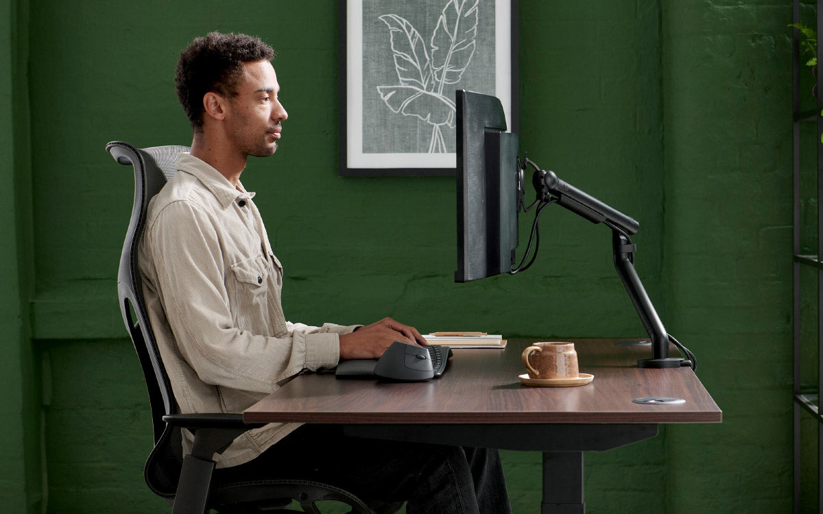 A man sat in a Herman Miller Cosm at a Jarvis desk with a Flo monitor arm as part of an ergonomic home office.