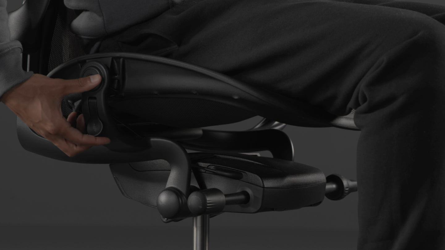 A video of a person sitting in a black Aeron gaming chair by Herman Miller, reaching beside the chair to adjust the seat height.