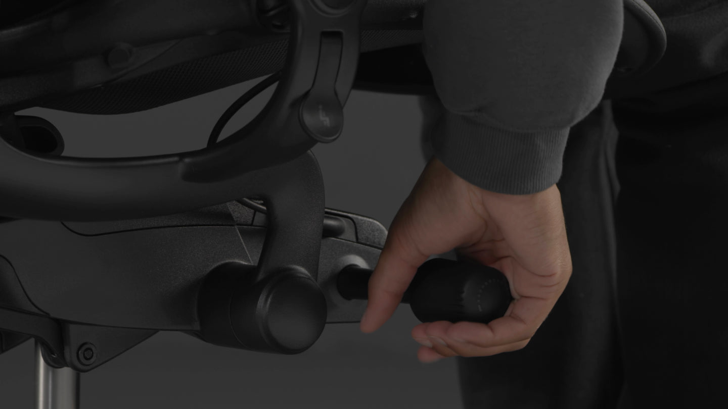 A video of a person sitting in a black Aeron gaming chair by Herman Miller, reaching beside the chair to adjust the tilt tension.