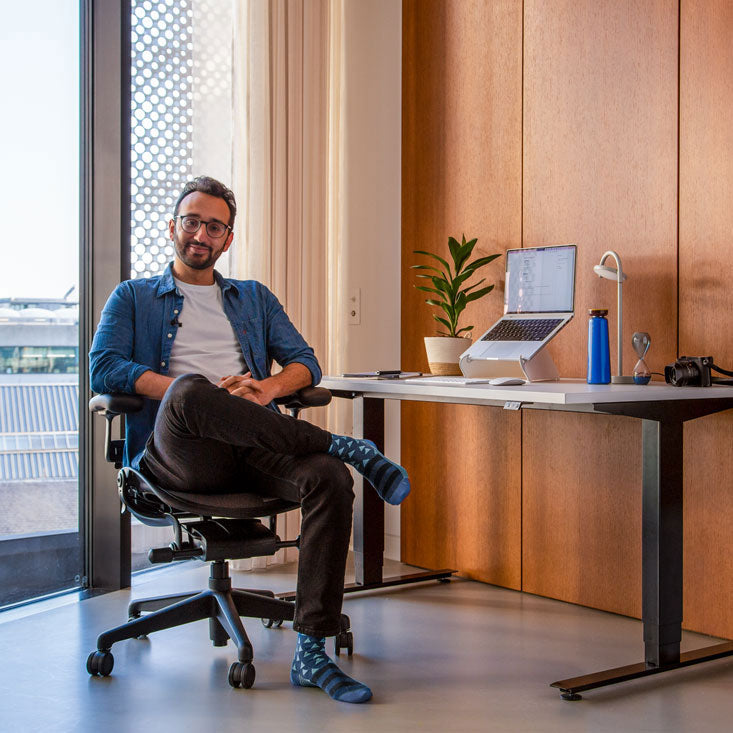 Photo of Ali Abdaal, podcaster, sat in Herman Miller Aeron office chair in graphite in front of Nevi sit-stand desk with black legs and white top with an laptop in an Oripura laptop stand
