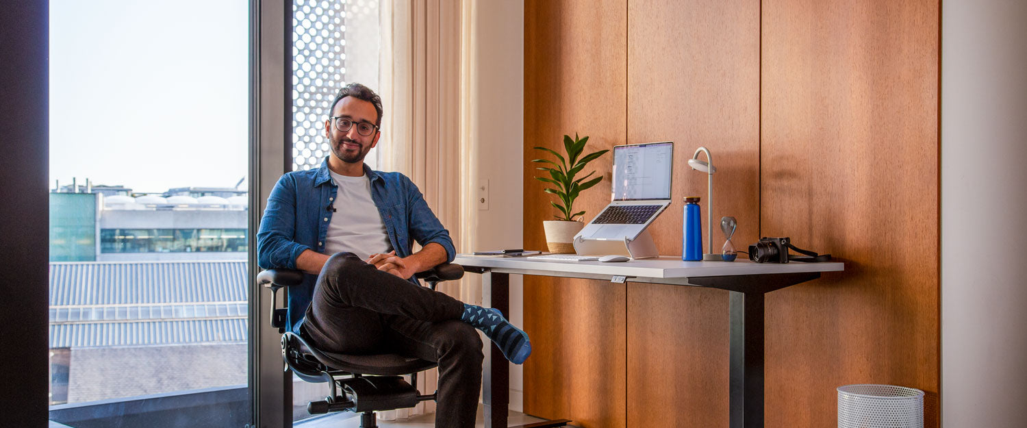 Photo of Ali Abdaal, podcaster, sat in Herman Miller Aeron office chair in graphite in front of Nevi sit-stand desk with black legs and white top with an laptop in an Oripura laptop stand