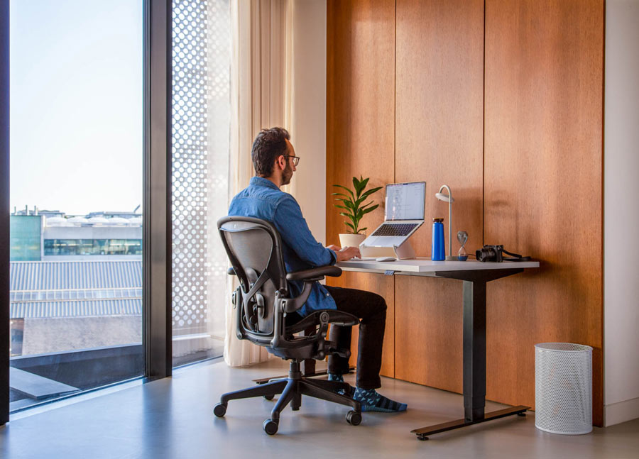 Photo of Ali Abdaal, podcaster, sat in Herman Miller Aeron office chair in graphite in front of Nevi sit-stand desk with black legs and white top with an laptop in an Oripura laptop stand