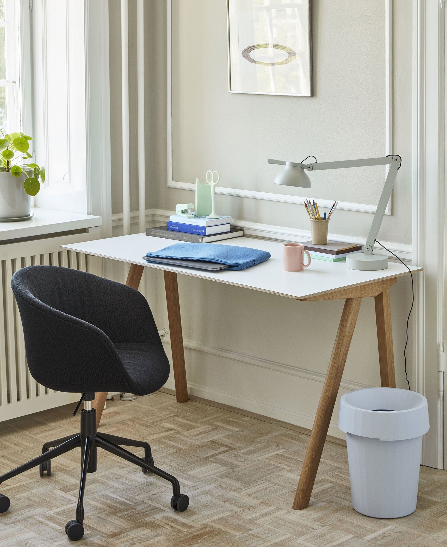Simple photo in home setting with view of garden of Hay Copenhague 90 desk in oak with white top, with Hay Slant lamp in khaki green on top and Hay small storage crate on the floor