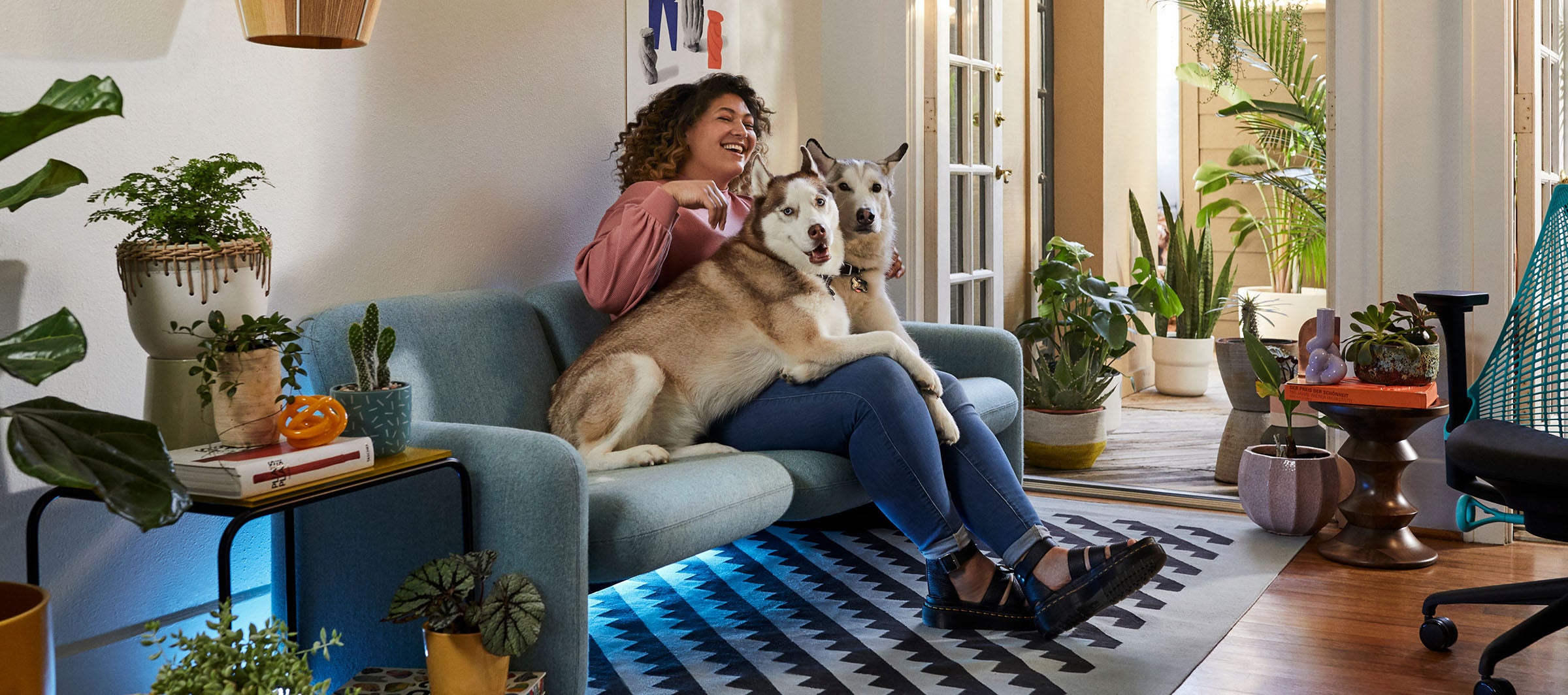 Friskk sat on Wilkes sofa in green lauching with her two dogs, Sayl gaming chair in black and ocean blue to the side