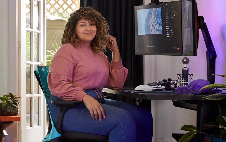 Pro streamer and voice actor Friskk sat on Herman Miller Sayl gaming chair in Ocean deep blue and black, at a black Nevi sit-stand gaming desk with monitor on Oliin monitor arm.