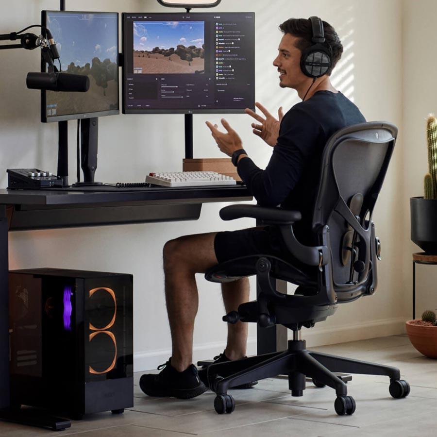Gamer and streamer Xaryu in a gaming set up sat on Herman Miller Aeron gaming chair with black Nevi sit-stand gaming desk and dual monitors on Ollin moniter arms