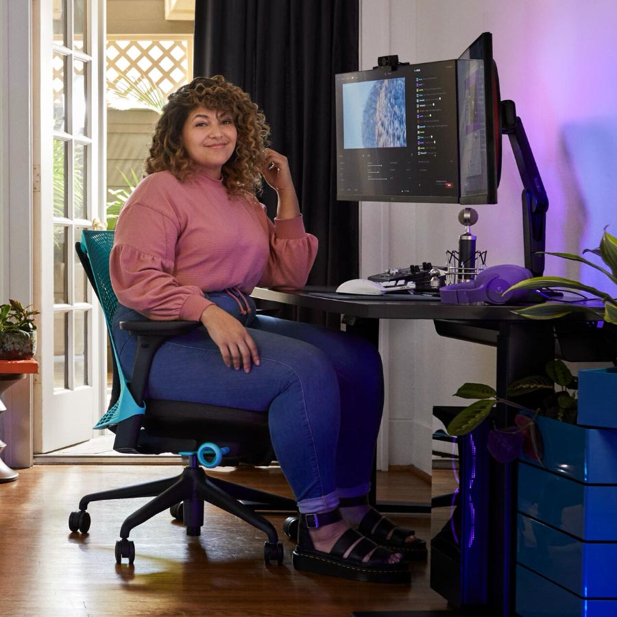 Pro streamer and voice actor Friskk sat on Herman Miller Sayl gaming chair in Ocean deep blue and black, at a black Nevi sit-stand gaming desk with monitor on Oliin monitor arm.