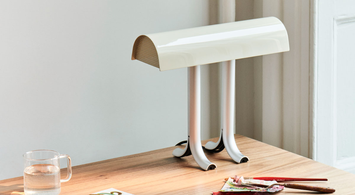 A cream and silver Anagram table lamp by HAY, positioned in the back corner of a wooden desk.