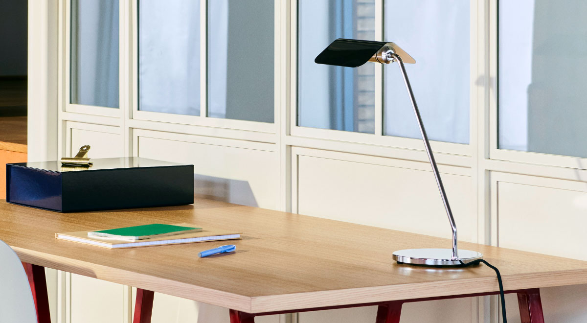 A black and silver Apex desk lamp by HAY, positioned in the back corner of a wooden desk.