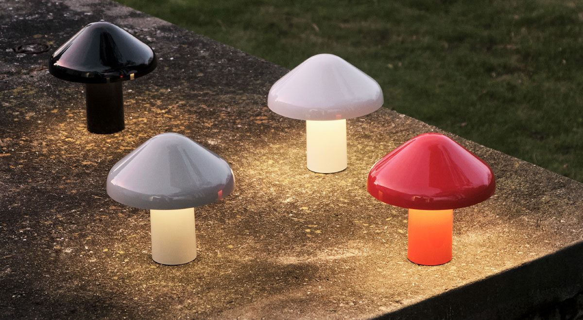 Four Pao portable table lamps by HAY in black, white, grey and red. Set outdoors in a garden, emitting a soft warm glow.