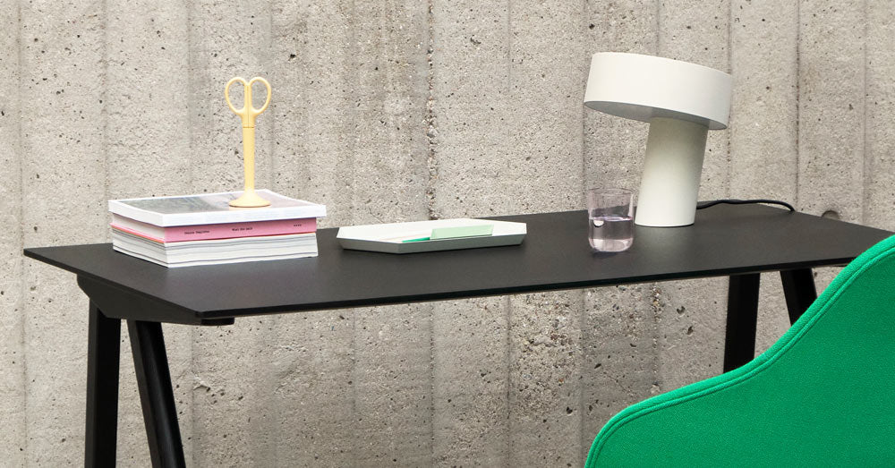 A white Slant lamp by HAY, positioned on a black Copenhague 90 desk by HAY, alongside a stack of books, yellow scissors and a water glass.