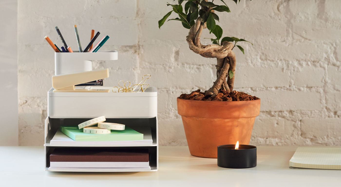 A white Formwork paper tray, stationary box and pen cup by Herman Miller, sitting on a white desktop with an indoor plant and a candle.