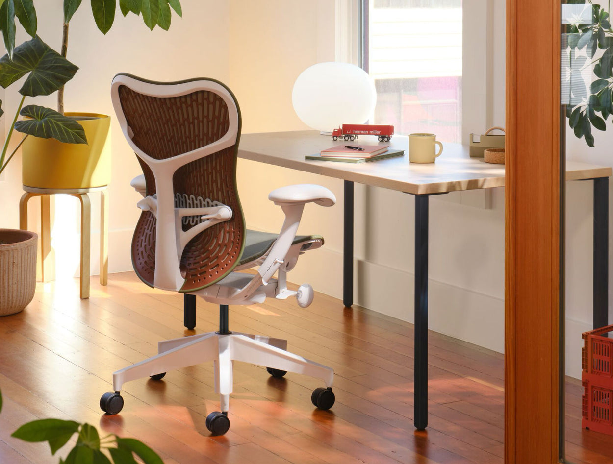 An olive green and white Mirra 2 office chair with a maroon back, positioned infront of a Herman Miller OE1 desk. Set in a home office environment, with solid wood flooring, white walls and warm, cosy lighting.