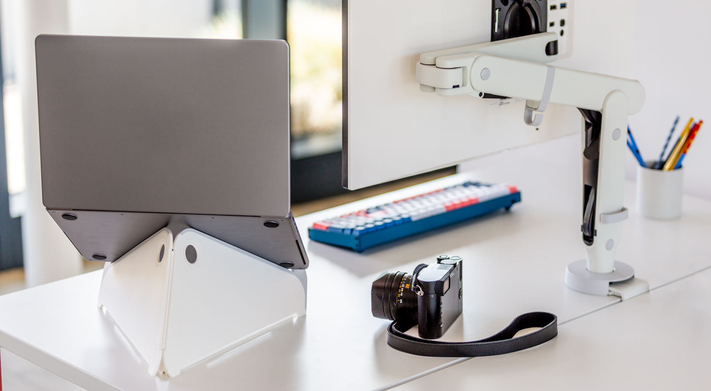 A white Oripura laptop stand by Herman Miller holding a laptop alongside a white monitor arm.
