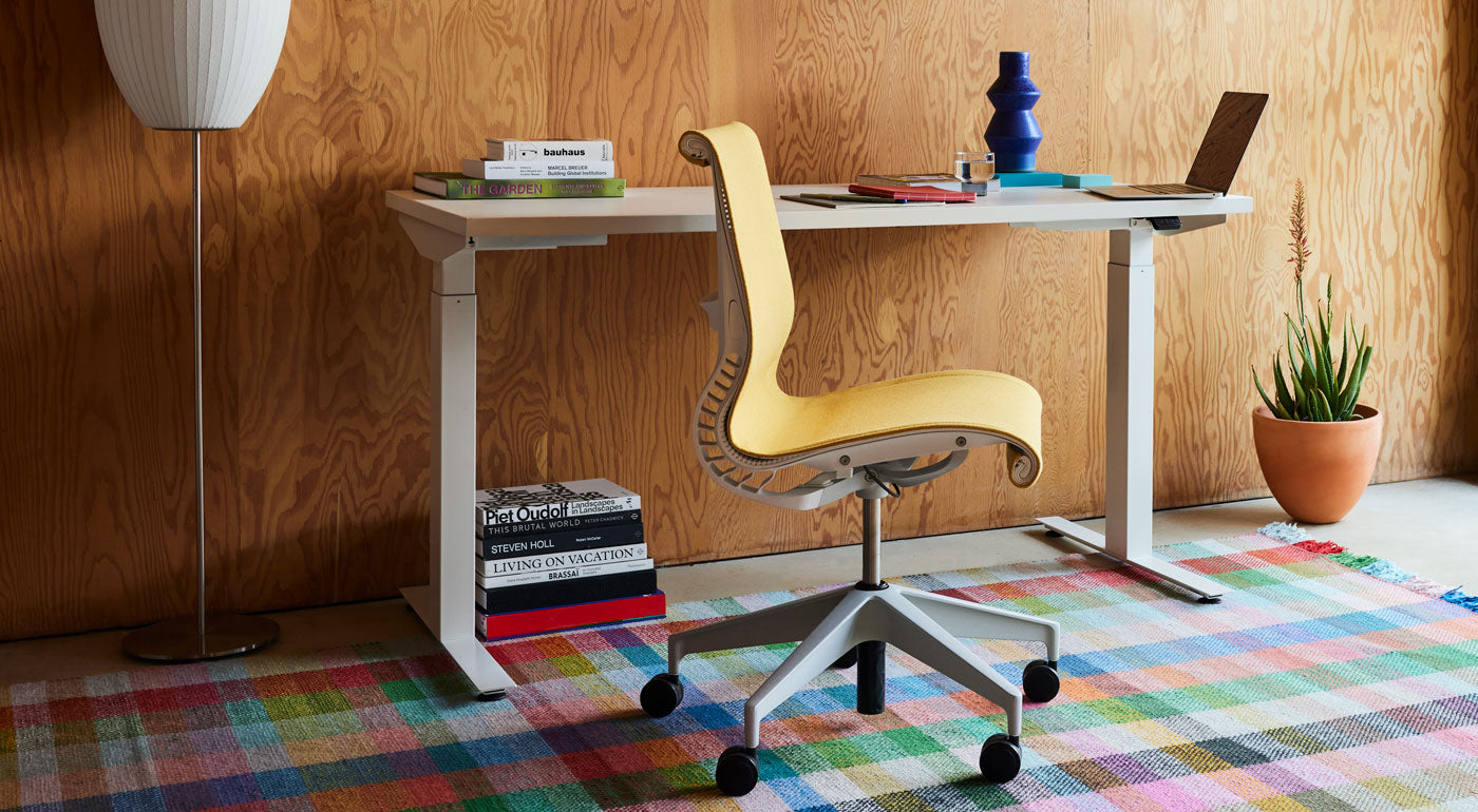 A yellow Setu office chair by Herman Miller, upholstered in Mimosa Meld fabric, positioned in front of a white Nevi standing desk on a colourful checked rug.