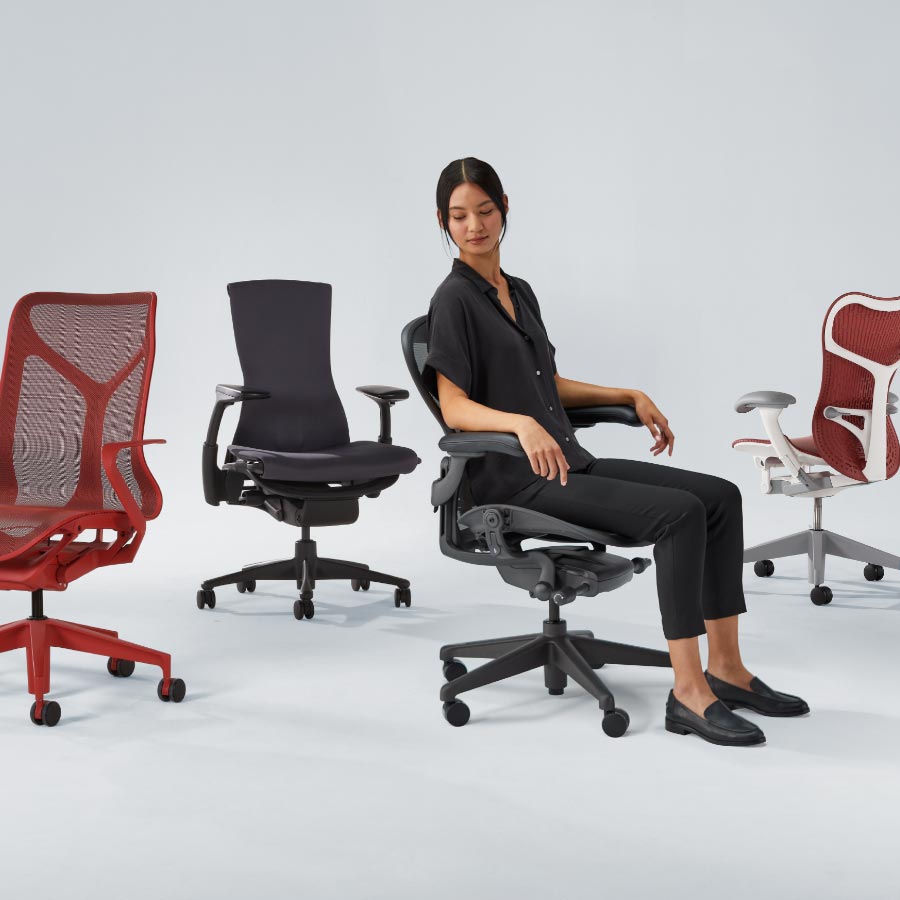 Line up of four Herman Miller office chairs including Cosm mid back in canyon red, Embody in black, Aeron in graphite with lady sat on and finally Mirra 2 with white frame and red seat and back