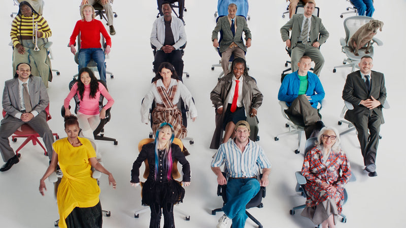 A large, diverse group of people sat on a selection of Herman Miller ergonomic office chairs in a white studio environment.