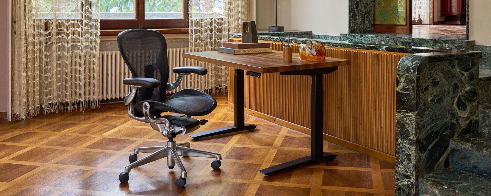 An Onyx black and polished aluminium Herman Miller Aeron alongside a walnut-topped Jarvis sit-stand desk in a mediterranean home office featuring wood paneling and black emerald marble.