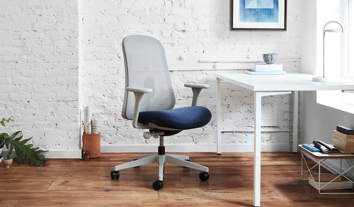 A grey and blue Lino office chair by Herman Miller, is positioned in front of a white desk. The interior space features a white brick wall, wooden flooring and a white Marselis table lamp on the desk.