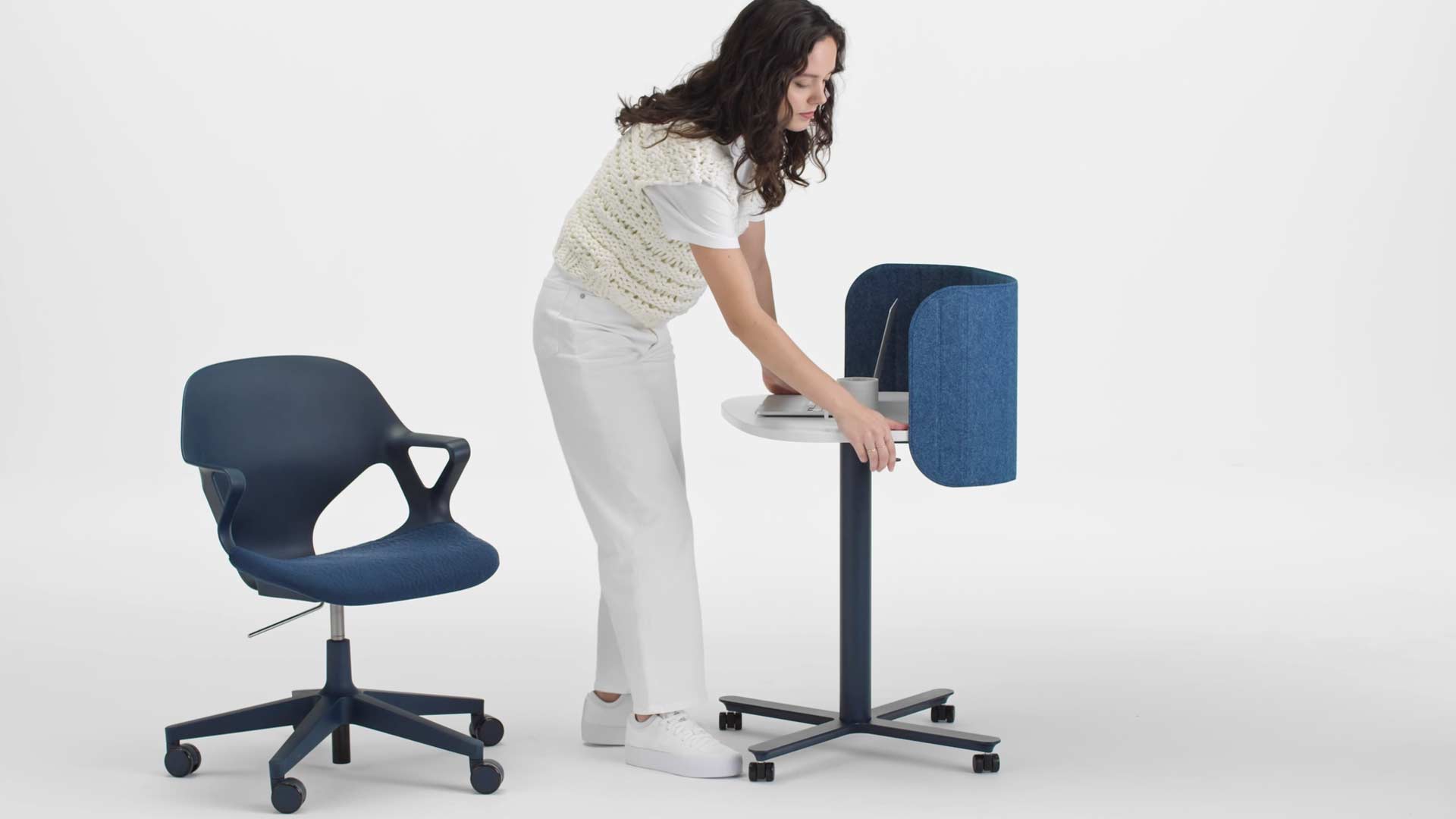 A video of a woman sitting in a blue Zeph office chair standing up and raising the height of a blue and white Passport work table by Herman Miller, using the lever on the side.