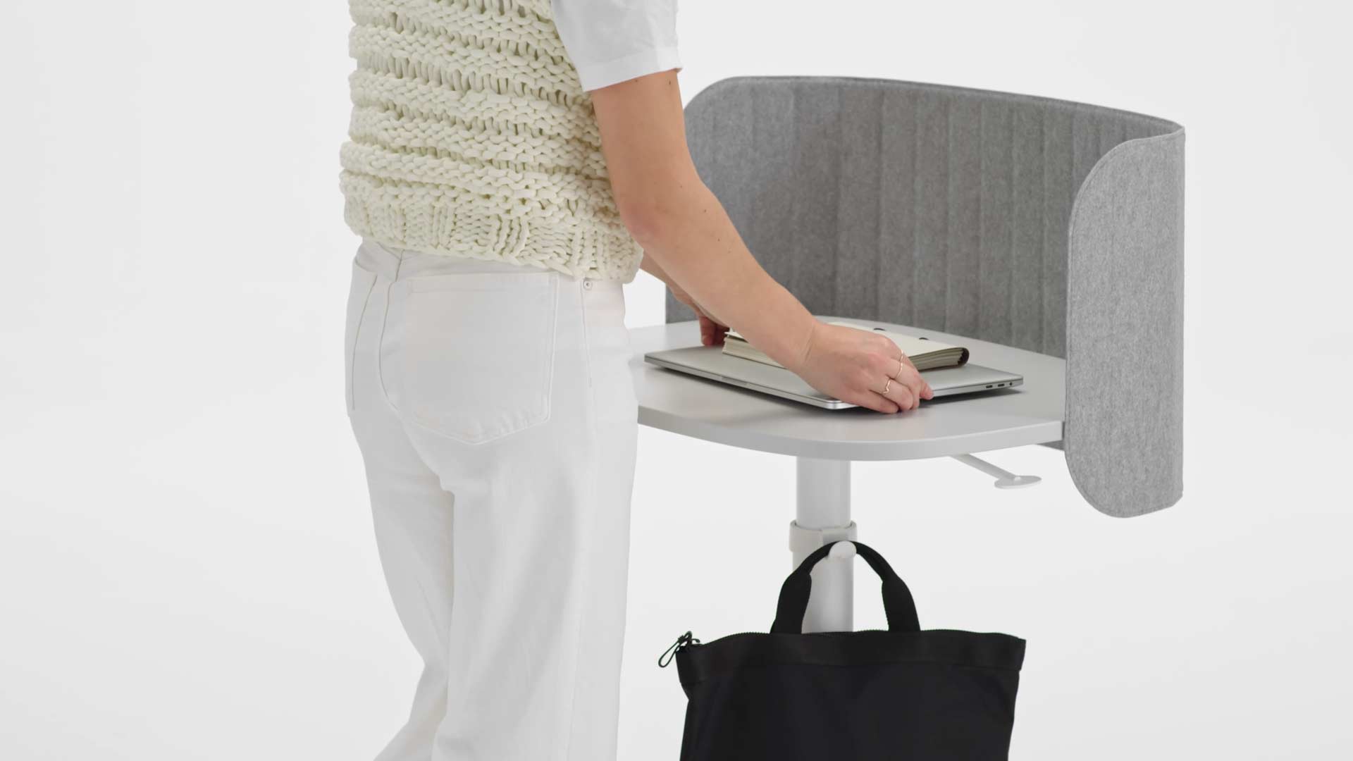 A video of a woman using a white Passport work table. She walks up to the desk, hangs her bag on the bag hook, places her laptop on the tabletop and then raises the desk to the correct height.