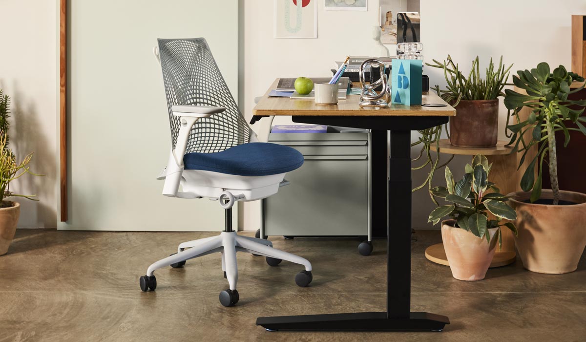 A white and blue Sayl office chair by Herman Miller, positioned next to a Jarvis standing desk with black legs and a bamboo worktop. Set in a home office environment, with a blue OE1 trolley and a selection of indoor plants in terracotta pots.